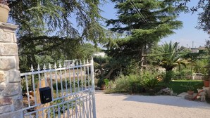 Une entrée fermée par un portail donne sur une allée de gravier qui mène à un jardin luxuriant, où l’on trouve une variété de plantes et d’arbres.