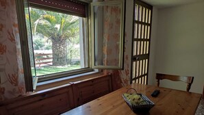 Une table de salle à manger en bois, sur laquelle se trouve un panier de collations et une télécommande. On aperçoit une fenêtre donnant sur un palmier et une clôture.