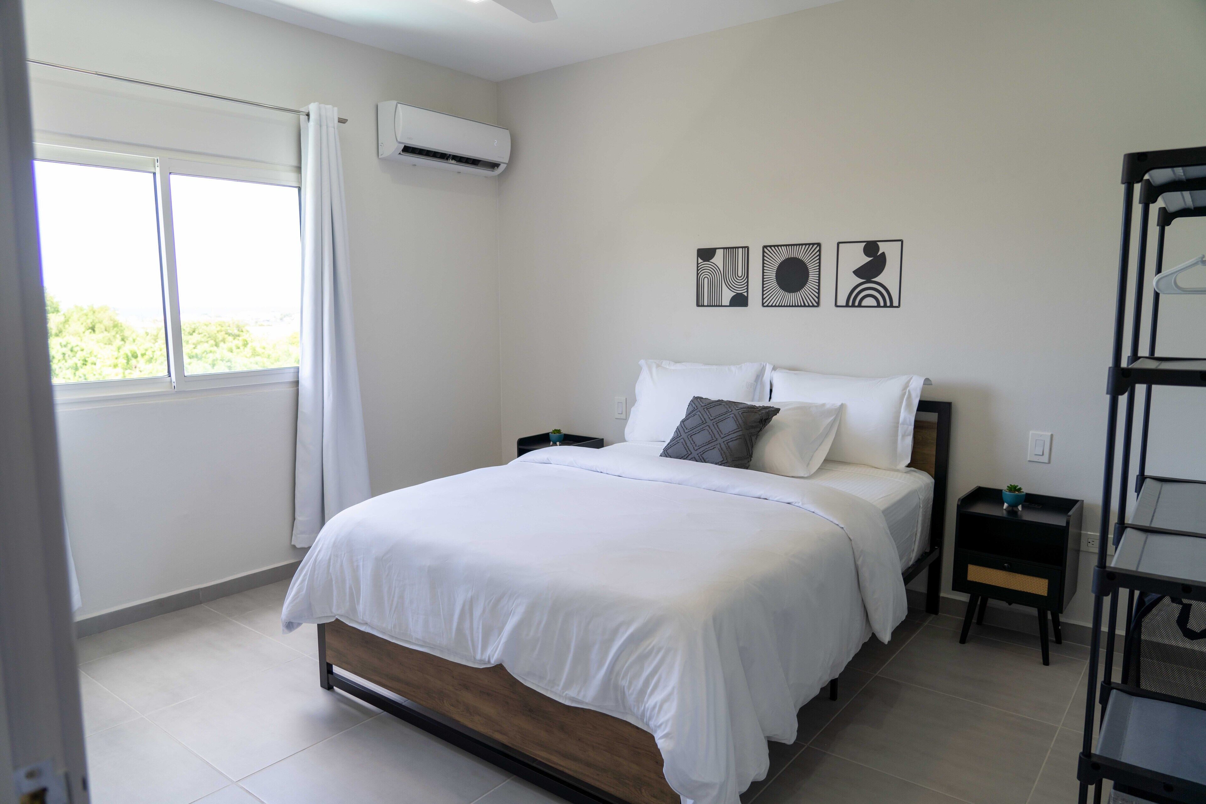 A bedroom with a bed, a nightstand, a wall-mounted air conditioner, and a window with curtains.