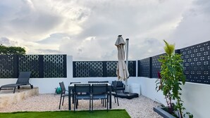 Una terraza en la azotea con mesa y sillas, una silla de descanso, un pequeño jardín y una cerca decorativa.