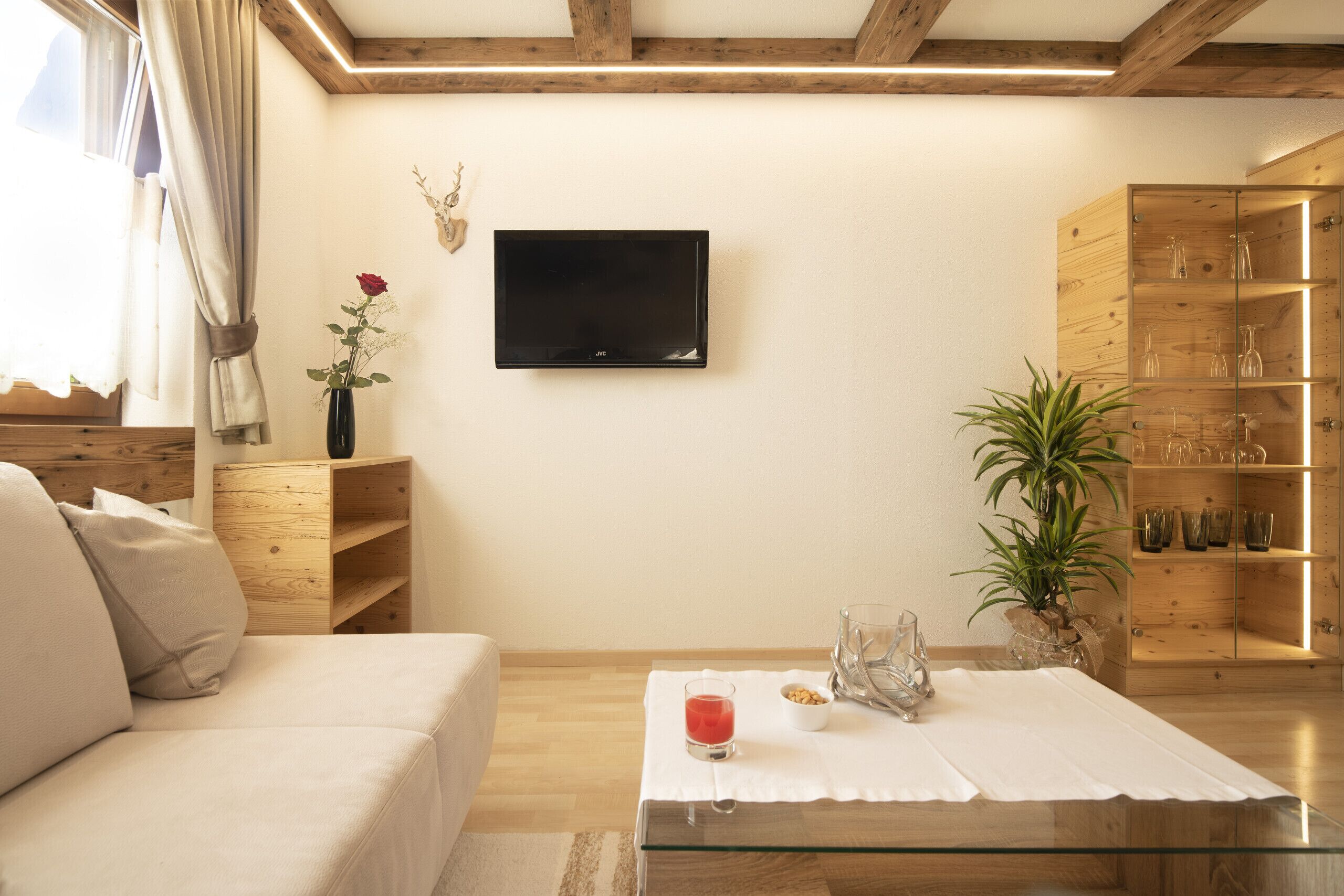 Una sala de estar moderna con un televisor de pantalla plana montado en la pared, una mesa de centro de vidrio, un sofá color beige, estanterías de madera con cristalería y una planta en maceta.