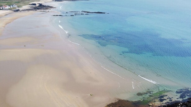 Vista aérea de uma área costeira com uma praia arenosa, águas azul-claras e montanhas ao longe.