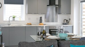 Cozinha moderna com armários cinzas, coifa de aço inoxidável e área de jantar com mesa de vidro posta para dois.