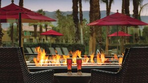 A fire feature with two glasses of beer in front of it, surrounded by outdoor seating with red umbrellas.