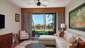 A living room with a sofa, armchair, coffee table, and a television. There is a large window with curtains, a ceiling fan, and a painting on the wall.