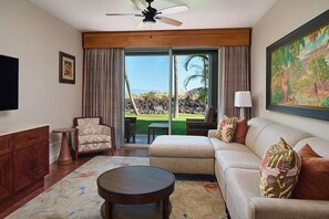 Ein Wohnzimmer mit Sofa, Sessel, Couchtisch und Fernseher. Ein großes Fenster mit Vorhängen, ein Deckenventilator und ein Bild an der Wand sind ebenfalls vorhanden.