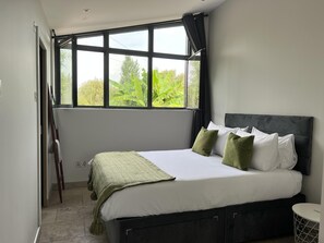 A bedroom with a large bed, a window with a view of greenery, and a bedside table. - River View Guest House, Jacuzzi, Boat, Fire Hire (Staines)
