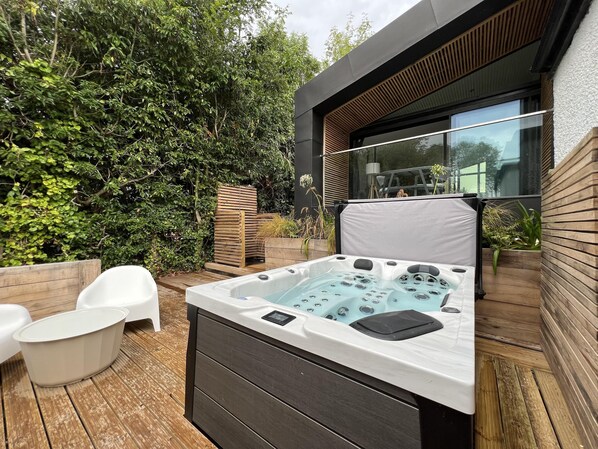 A modern outdoor hot tub area with a wooden deck, white chairs, and a lush green hedge backdrop. - River View Guest House, Jacuzzi, Boat, Fire Hire (Staines)