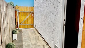 A narrow outdoor corridor with a concrete floor, a wooden fence on one side, and a white textured wall on the other. There are two potted plants and a door leading to a dark interior. - Studio Suite with Private, Secured Entrance, No Shared Spaces, Driveway Parking  (Spring Valley)