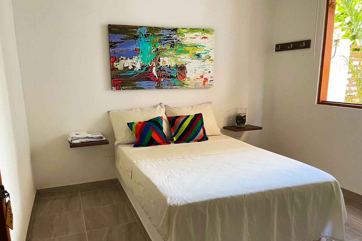Un lit bien fait, avec une literie blanche et des coussins rayés aux couleurs vives, une petite table de chevet avec un verre d’eau, et une peinture abstraite aux couleurs vives accrochée au mur.