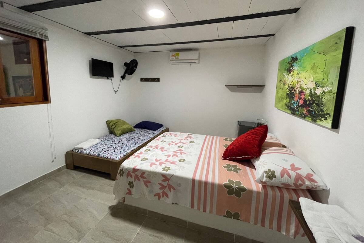 Une petite chambre bien éclairée, avec un lit simple, un petit lit, une télévision et un tableau au mur.