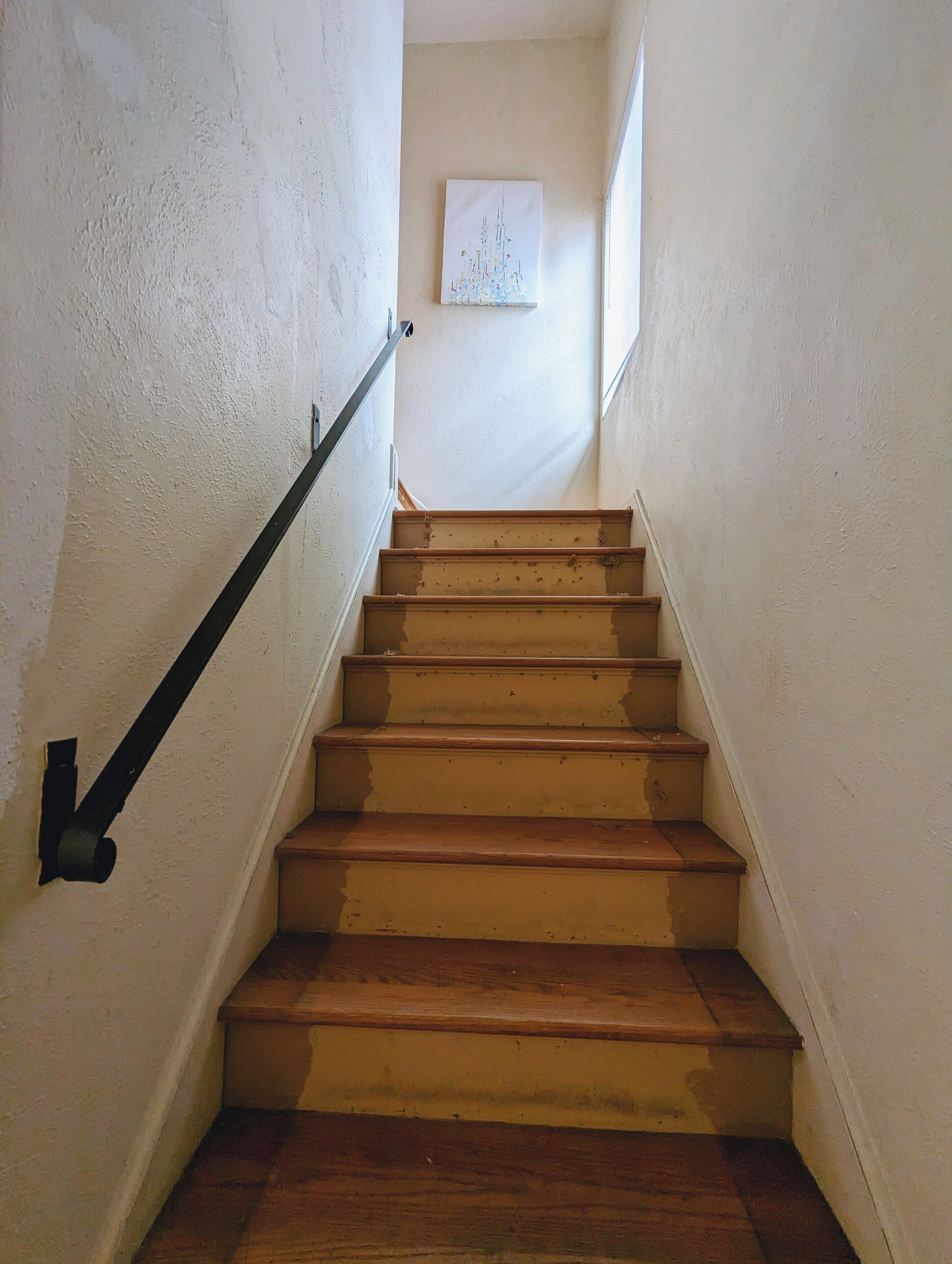 Un escalier avec des marches en bois et une rampe noire, menant à un palier où est accrochée une image encadrée au mur.