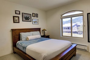 A bedroom with a wooden bed, a nightstand, a lamp, and a window with a view of a house and trees. - Queen Bee - Beach Front w/ Vast Ocean Views, Spa (Gold Beach)