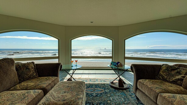 Quarto com três amplas janelas com vista para o oceano, uma pequena mesa com uma bebida e sofás estampados.