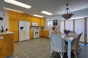 A kitchen with wooden cabinets, a white refrigerator, a microwave, an oven, and a dining area with wicker chairs and a table. - Queen Bee - Beach Front w/ Vast Ocean Views, Spa (Gold Beach)