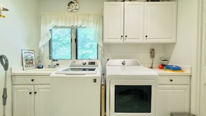 A laundry room with a front-loading washer and dryer, a window with a curtain, and a small trash bin. - Peaceful, Woodland Retreat in Frankfort, MI Near Crystal Lake & Sleeping Bear (Frankfort)