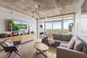 A modern living room with a large TV, a sofa, a coffee table, and a view of a cityscape through large windows. - 3250 Ne Apartament - Wynwood Walkable (Miami)
