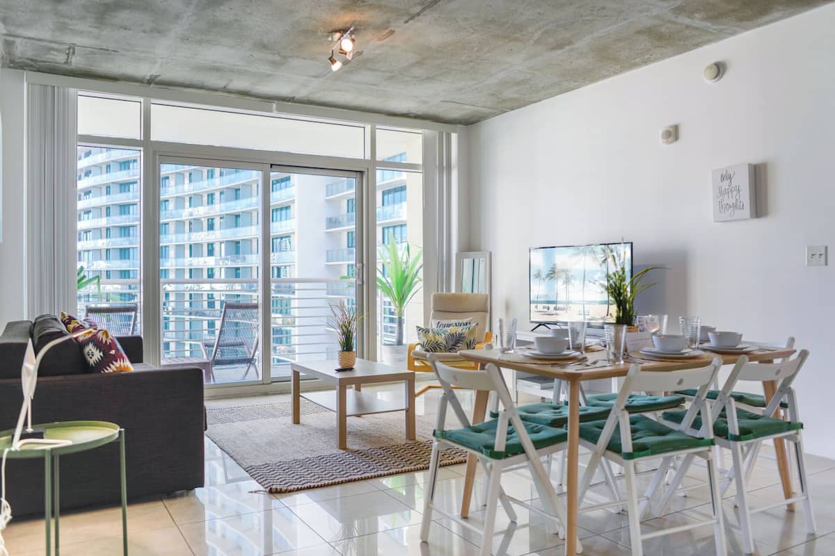 A modern living room with a large window, a dining area with a TV, and a balcony with a view of high-rise buildings.