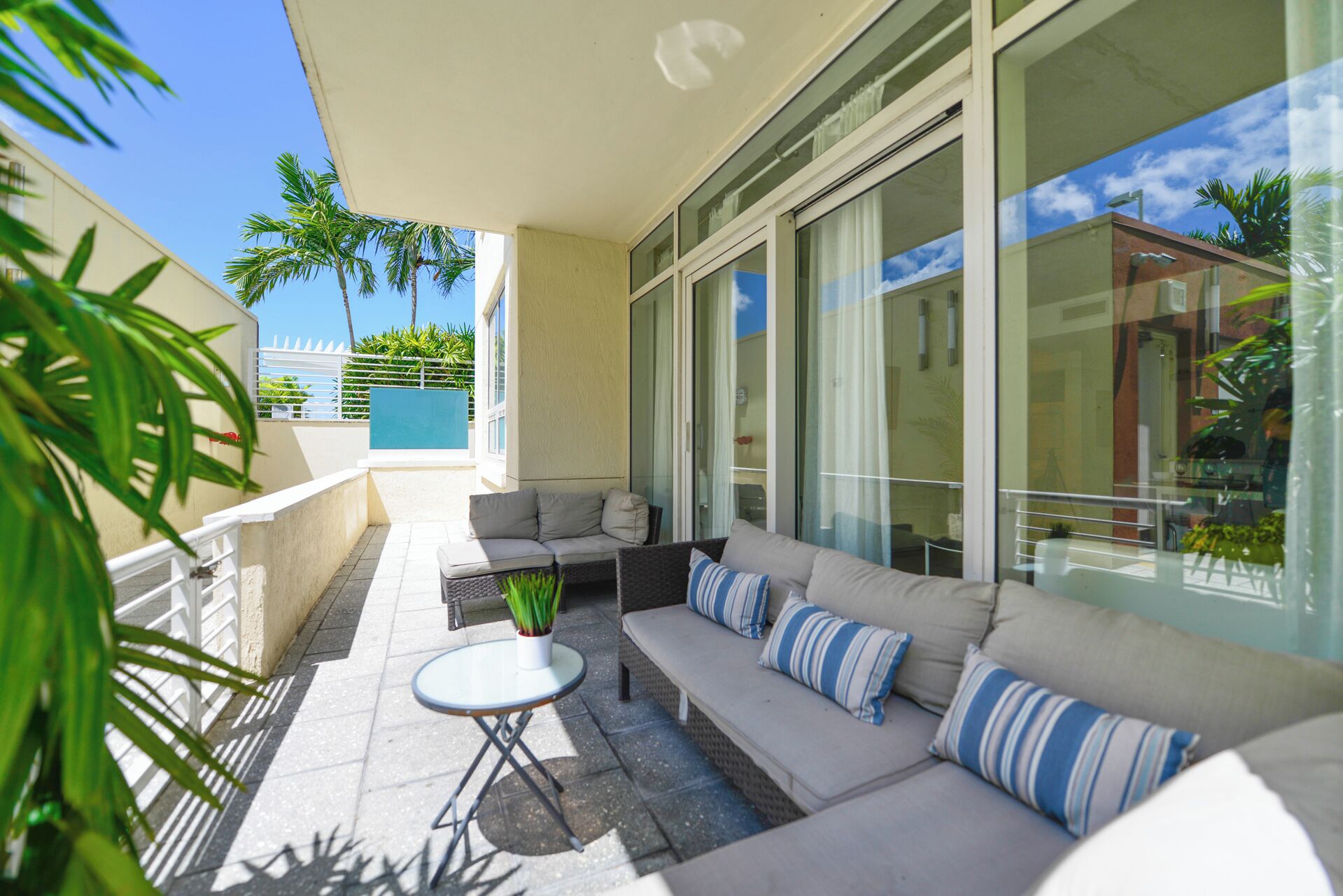 A sunlit outdoor seating area with a sofa and a small table, surrounded by large windows and a view of palm trees.