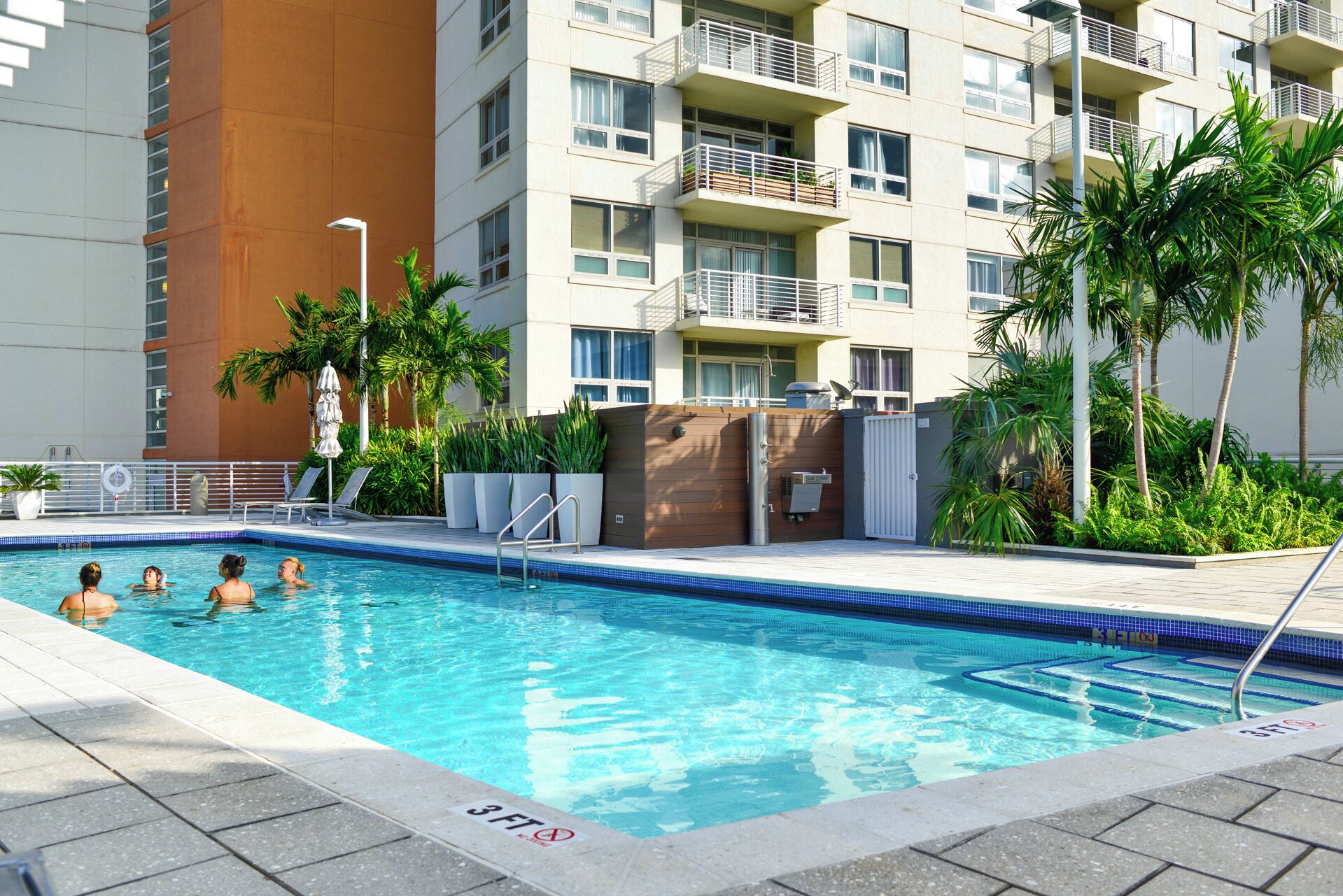 A swimming pool with a 3 FT depth limit sign, surrounded by palm trees and a multi-story building with balconies.