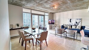 Un salon moderne avec une table à manger en verre, des sièges confortables, un grand téléviseur et une vue sur la ville grâce à de grandes fenêtres.