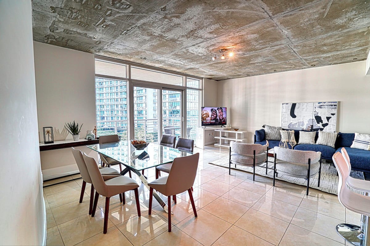 A modern living room with a glass dining table, comfortable seating, a large TV, and a city view through large windows.