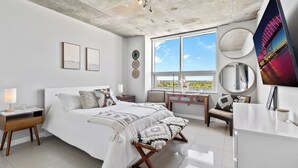 Une chambre à coucher moderne, dotée d’un grand lit, de tables de chevet, d’un bureau et offrant une vue sur le paysage urbain grâce à de grandes fenêtres.