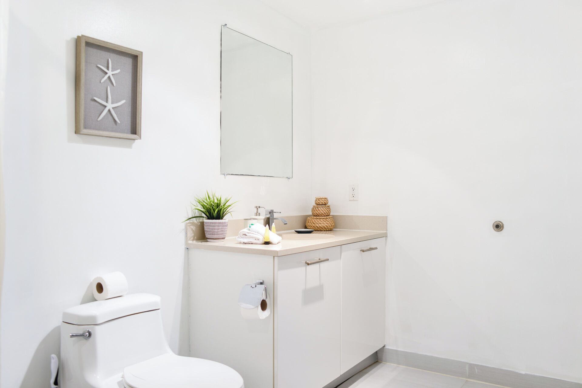 A bathroom with a toilet, sink, and mirror. There is a framed picture on the wall and a plant on the counter.