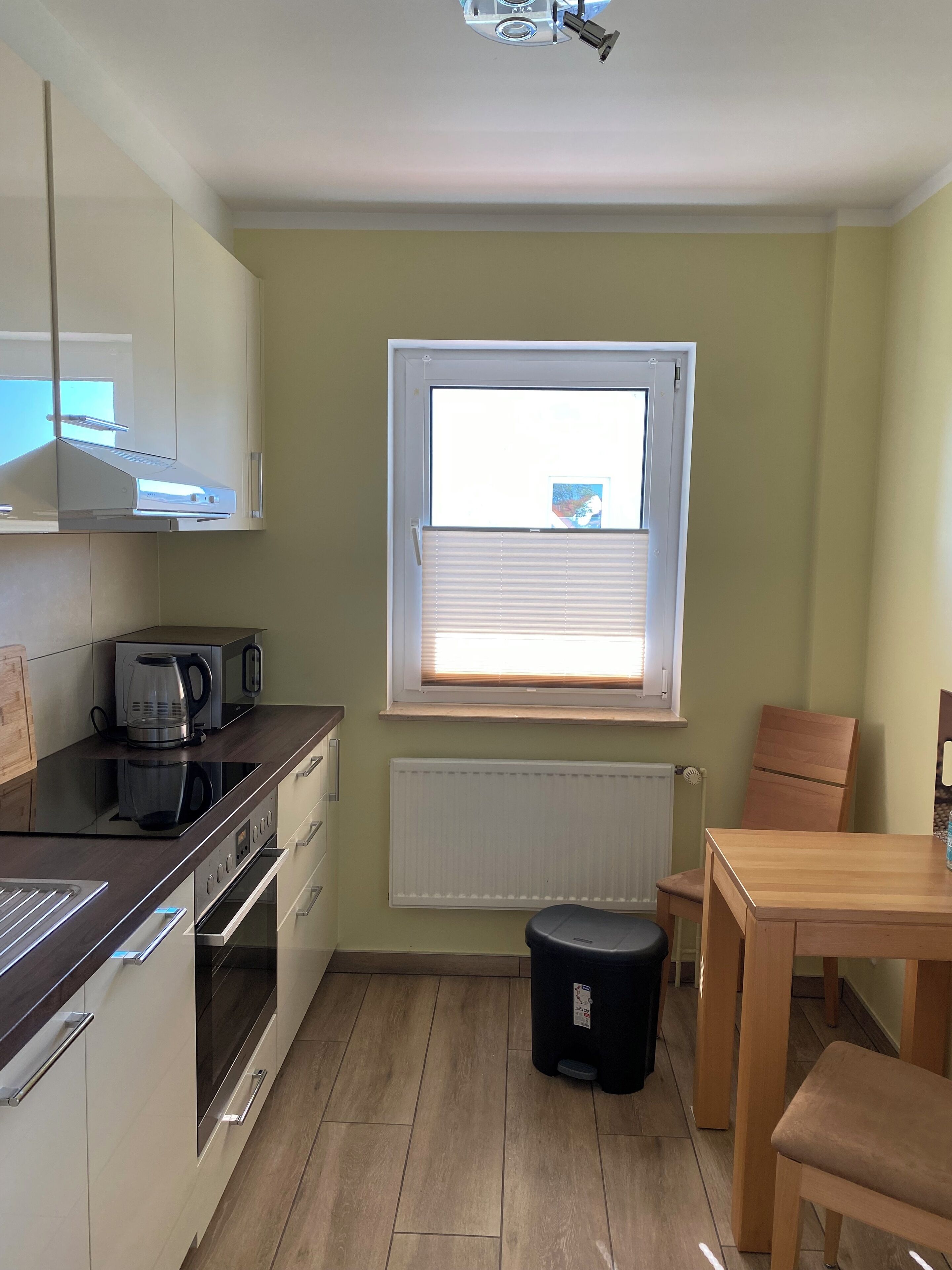 A compact kitchen with a coffee maker, a kettle, and a toaster on the counter. There is a window with blinds, a radiator below, and a small stool near the table.