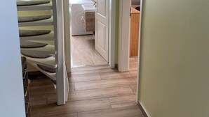 Un couloir avec un plancher en bois, une porte blanche sur la gauche et une fenêtre avec des stores.