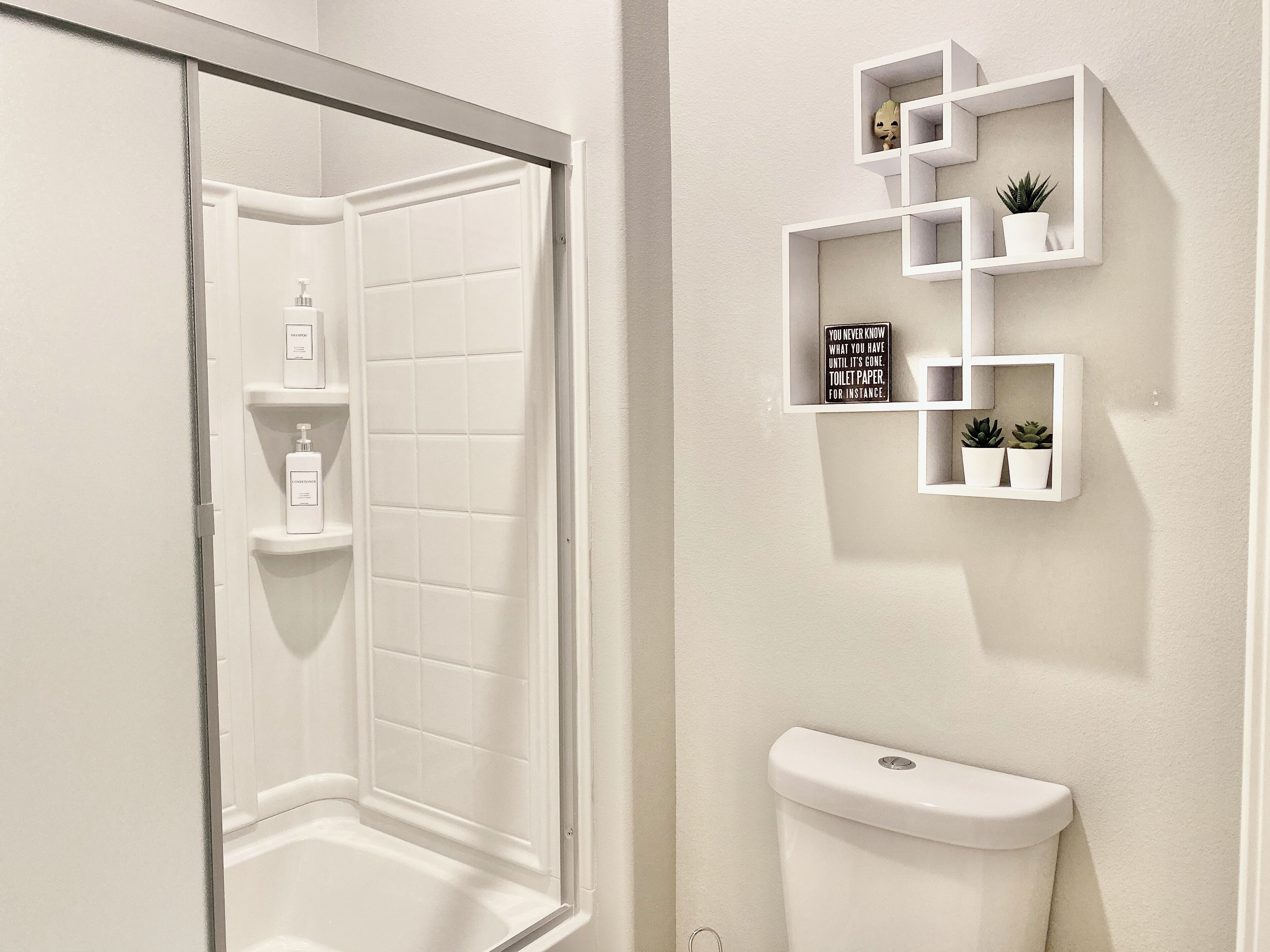 Een badkamer met een douche, toilet en wandplanken met toiletartikelen en een grappig bord.