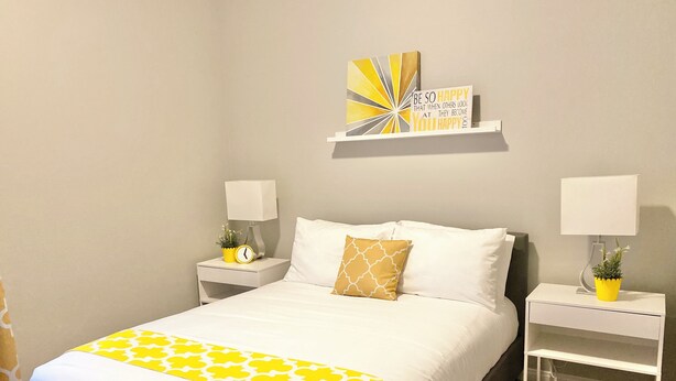A hotel room with a bed, two bedside tables, a nightstand, a wall clock, a decorative piece, and a shelf with a motivational quote.