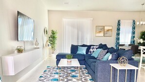 A modern living room with a sectional sofa, a flat-screen TV, a dining area, and a large window with curtains. - Desert Poolside Haven (Cathedral City)