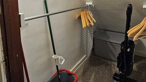 Une petite pièce, recouverte de moquette, où un balai, une pelle et une serpillière sont appuyés contre le mur.