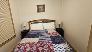 Un lit bien fait, recouvert d’une courtepointe à motifs, deux tables de chevet avec des lampes, un tableau encadré au mur et une fenêtre avec des stores.