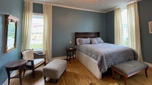 A bedroom with a bed, a chair, an ottoman, a nightstand, and a mirror.