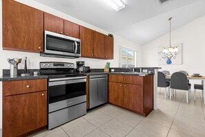 A modern kitchen with wooden cabinets, stainless steel appliances, a dining area with a chandelier, and a window. - Disney Area 4BR Home with Heated Private Pool (Kissimmee)