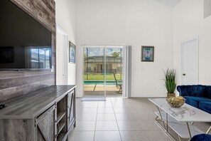 A spacious living area with a sliding glass door leading to a backyard, a blue sofa, and a marble coffee table. - Disney Area 4BR Home with Heated Private Pool (Kissimmee)