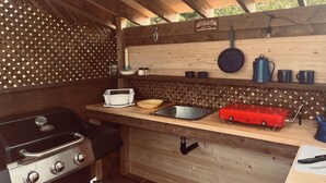 Een houten buitenkeuken met een barbecue, spoelbak en diverse kookgerei.