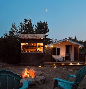 Un espace extérieur accueillant avec un foyer, deux chaises turquoise et une petite structure en bois décorée de guirlandes lumineuses.