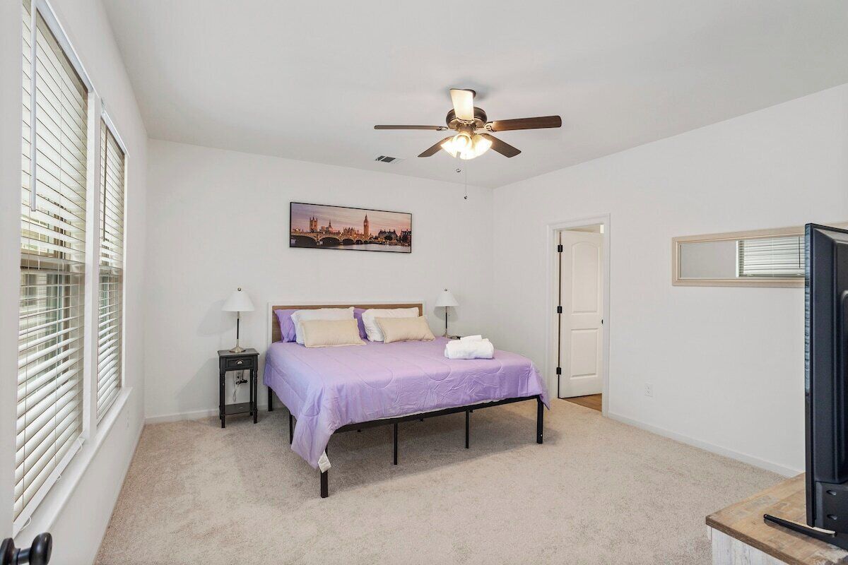 A bedroom with a bed, a ceiling fan, a nightstand, a lamp, a door, and a window with blinds.