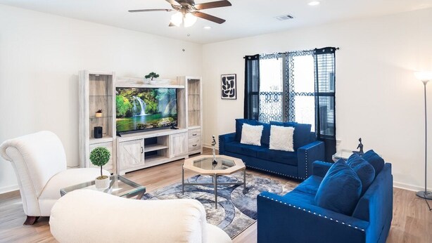 Una sala de estar con un sofá azul, un sillón blanco, una mesa de centro de vidrio, un televisor sobre un mueble de madera y un ventilador de techo.