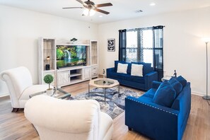 Een woonkamer met een blauwe bank, een witte fauteuil, een glazen salontafel, een tv op een houten dressoir en een plafondventilator.