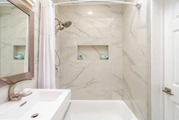Un baño moderno con un lavamanos blanco, un espejo y una zona de ducha con paredes de mármol y un cabezal de ducha.