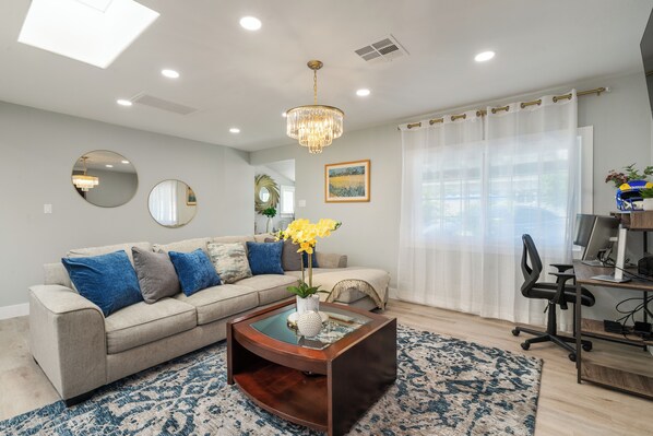 A living room with a beige sofa, a glass coffee table, a chandelier, and a large window with sheer curtains.