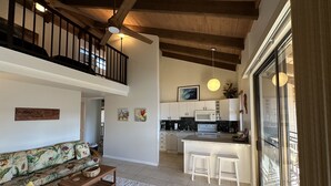 A spacious living area with a high ceiling, wooden beams, a ceiling fan, a sofa with floral upholstery, a coffee table, and a kitchen area with white cabinets and a large window.