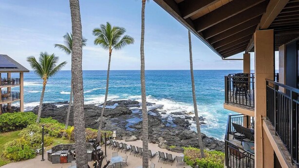 A beachfront property with a balcony overlooking the ocean, palm trees, and a rocky shoreline.
