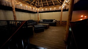 Een houten terras met rieten dak, voorzien van banken en een tafel, en een trap die omhoog leidt.