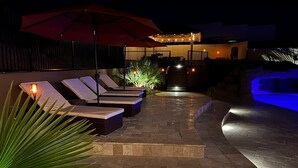 Een terras buiten bij nacht, met ligstoelen, rode parasols en een zwembad met blauwe verlichting.