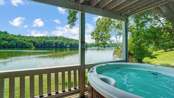 A hot tub on a covered deck with a view of a lake and forest. - Lakefront Home: Fishing + Kayaking + King Bed + Hot Tub (Logan)